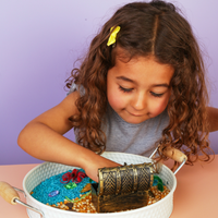 Young girl playing with a sensory play activity with a treasure chest and sea-themed rice in a white container against a purple background.