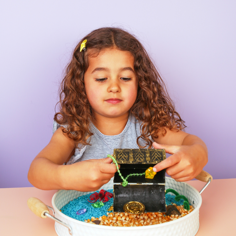 Child playing with a small treasure chest in a sensory play tray filled with rice, pebbles, and toys, perfect for screen-free play ideas for young kids.