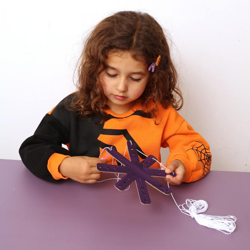 Child wearing an orange and black outfit with spider web designs, sitting at a table with a purple craft project from My Mini Maker Halloween craft kit for kids 