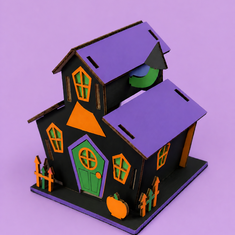 Finished haunted house project from the Halloween Project Kit, a wooden house painted black with a purple roof, orange and green windows, a green door, picket fence and a pumpkin by the step