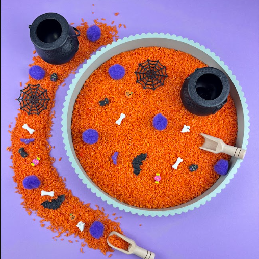 Halloween sensory tray filled with orange rice, purple pom-poms, small bones, bats, spider webs, ghosts and two black cauldrons, with a wooden scoop