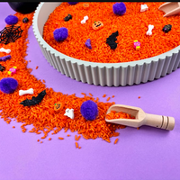 Close-up of the Halloween sensory tray: orange rice spilling onto the table with purple pom-poms, a bat, a bone, a tiny ghost, a pumpkin and a wooden scoop