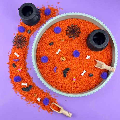 Halloween sensory tray filled with orange rice, purple pom-poms, bones, bats, spider webs and tiny ghosts, with two black cauldrons and a wooden scoop