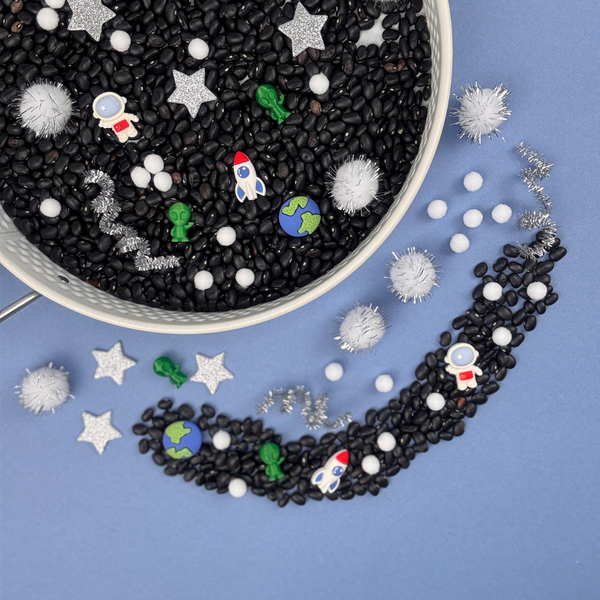 Space-themed sensory tray from My Mini Maker with black beans, colourful space-themed figurines, and a white bowl on a blue background.