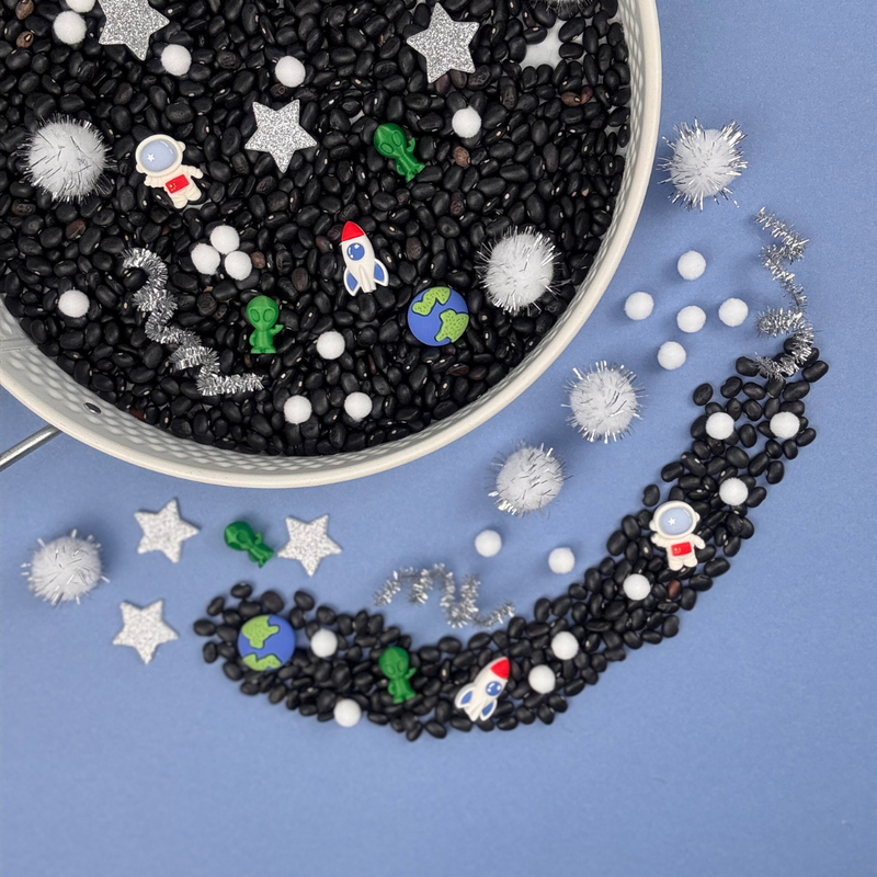 Space-themed sensory tray from My Mini Maker with black beans, colourful space-themed figurines, and a white bowl on a blue background.