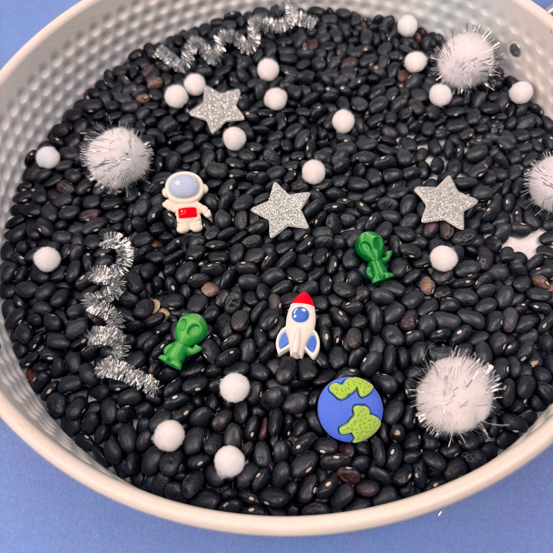 Space-themed sensory tray from My Mini Maker with black beans, stars, planets, and an astronaut figure.